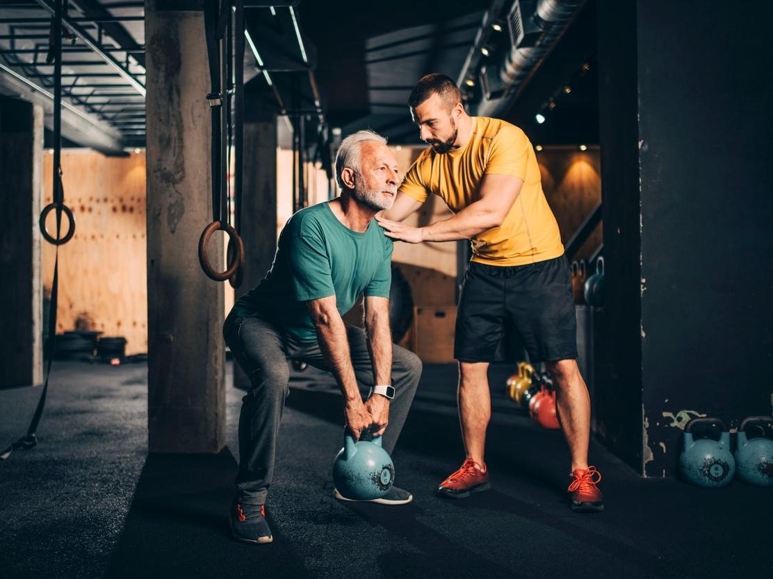 Hombre mayor levantando una kettlebell apoyado por un entrenador personal